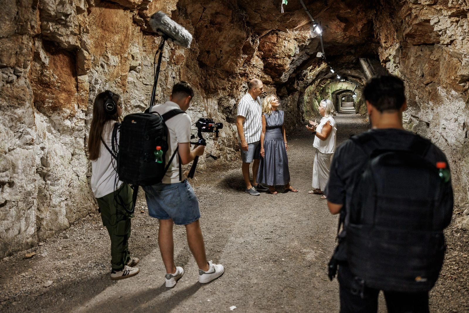 Video production crew filming three people talking inside a rock-carved tunnel
